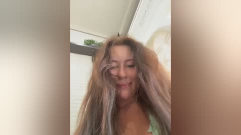 A blurry video of a woman with long, messy, light brown hair and a green top, taken indoors with a white wall and potted plant in the background.