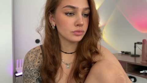 Video of a young woman with long, wavy brown hair, light skin, and full eyebrows. She wears a black choker, silver earrings, and a black top, with intricate tattoos on her left arm. Background includes a blurred, colorful abstract wall.