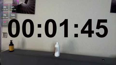 Video of a beige wall with a large black \"00:01:45\" clock, a bottle, and a framed art piece.