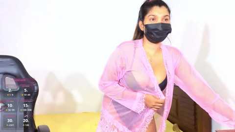 A video of a woman with medium skin tone, dark hair, and a black mask, wearing a sheer pink robe revealing a black bra. She stands in a room with a white wall and a yellow couch.