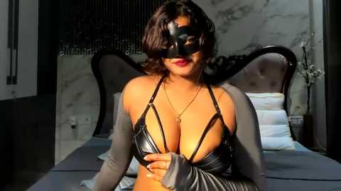 Media: Video of a woman with medium brown skin, dark wavy hair, wearing a black leather bra, black mask, and gray cardigan, seated on a bed with gray sheets and white pillows, marble wall in the background.