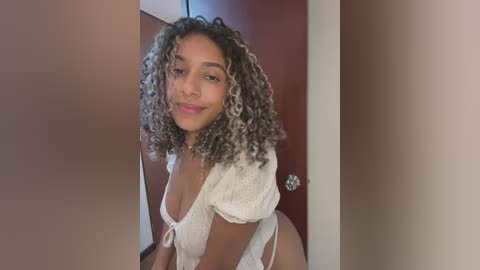 Video of a young, light-skinned woman with curly, highlighted hair, wearing a white lace top and white thong, posing confidently in a doorway.