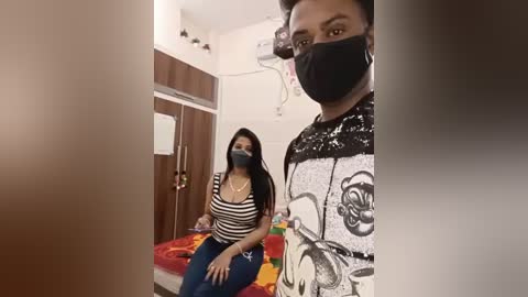 Video of a woman in a mask, black and white striped shirt, blue jeans, sitting on a bed in a small, sparsely furnished room with wooden panels and a fan.