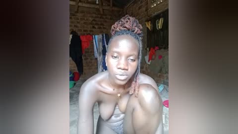 Video of a topless Black woman with braided hair, sitting in a rustic room with a brick wall, colorful clothes hanging, and a wooden floor.