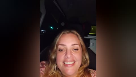 Video of a smiling, fair-skinned woman with long blonde hair, sitting in a dimly lit car, wearing a pink floral dress.