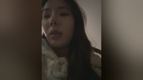 Media: A video of an East Asian woman with long, dark hair, wearing a cream-colored, fur-lined coat, taken in a dimly lit room, with shadows obscuring the background.