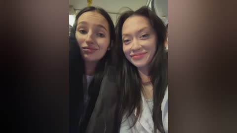 Video of two smiling young women with long black hair, wearing black jackets, taken indoors with blurred background.