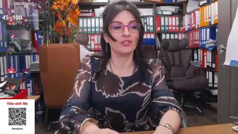 A video of a woman with glasses, black hair, and a patterned blouse, seated at a desk in a colorful, cluttered office with bookshelves and plants.