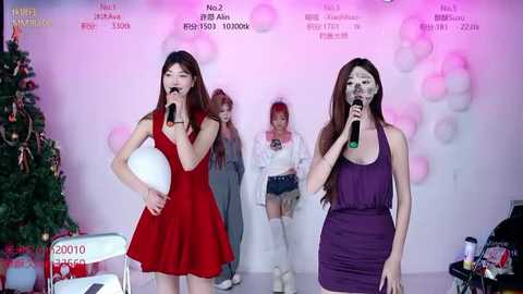 Video of two young East Asian women in sleeveless dresses, one red and one purple, singing into microphones, with a Christmas tree and pink balloons in the background.