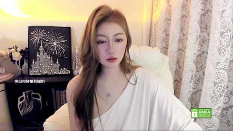 Media: A video of a fair-skinned woman with long, wavy brown hair, wearing a white off-shoulder dress, sitting indoors with a wall-mounted black art piece featuring fireworks in the background.