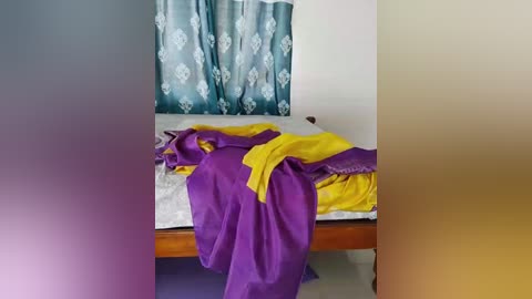 Video of a simple room with a bed covered in purple and yellow bed linens, a teal curtain with white floral pattern, and a plain white wall.