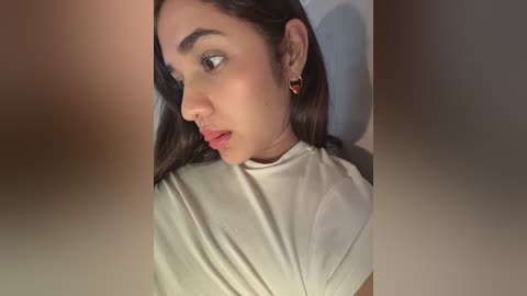 Media: A close-up video of a young South Asian woman with long, dark hair and medium skin tone, wearing a white turtleneck, sitting indoors.