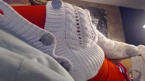 Video of a cozy room with a white, textured knit sweater draped over an orange couch. In the background, a person with short, light-colored hair is partially visible.