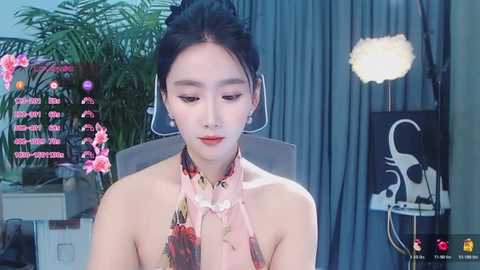 Media: Video of an Asian woman with fair skin, wearing a floral-patterned, off-shoulder top, standing indoors with greenery and a lamp in the background.