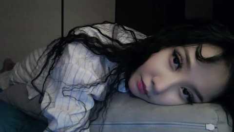 Video of a young Asian woman with wet, dark hair, lying on a gray couch, wearing a white and blue striped shirt, looking tired and contemplative.