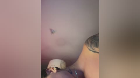 A blurry video of a topless, tattooed woman with fair skin, lying on her back in a dimly lit room. Her breasts are visible, and she wears a white cap.