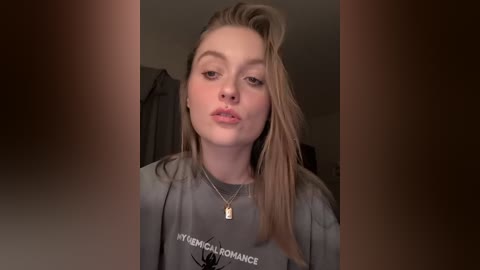 Video of a young woman with long, straight blonde hair and fair skin, wearing a grey t-shirt with \"YOUNG CALIFORNIA DANCE\" printed on it, indoors with blurred background.