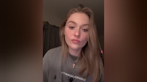 A video of a young woman with light skin and long blonde hair, wearing a grey hoodie with a black logo, sitting in a dimly lit room.