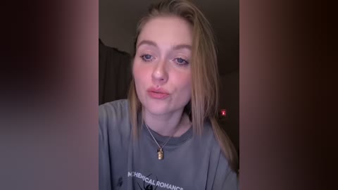 Video of a young Caucasian woman with light skin and long blonde hair, wearing a gray sweatshirt and a silver necklace. She's indoors with blurred background, giving a pouty expression.