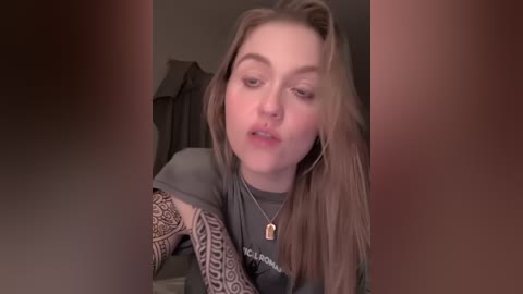 Video of a young woman with light skin and long, wavy brown hair, wearing a dark gray t-shirt and a dog tag necklace. She has intricate tattoos on her arms, captured in a dimly lit room with a blurred background.