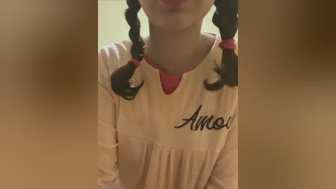 Video of a young girl with fair skin, wearing a beige dress with a pink collar and black braid hair accessories. She has a slight smile. Background is blurred, focusing on her face and shoulders.