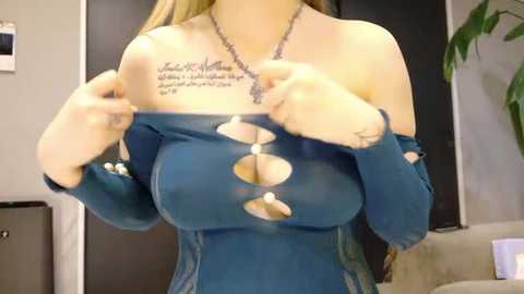 Media: Video of a fair-skinned woman with blonde hair, wearing a torn blue off-shoulder dress revealing cleavage and a tattoo on her chest. Background features a beige couch and a potted plant.