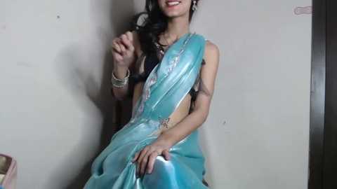 Video of a smiling South Asian woman in a turquoise sari with a black blouse, seated against a plain wall, wearing gold jewelry.