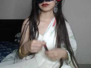 Video of an Indian woman with long black hair, wearing a white saree, gold bangles, and a black mask, adjusting her saree in a bedroom with a blue-patterned bedspread.