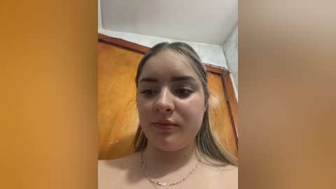 Video of a young woman with light skin, long blonde hair, and minimal makeup, standing in a bathroom with wooden walls and a beige shower curtain. She wears a silver necklace.