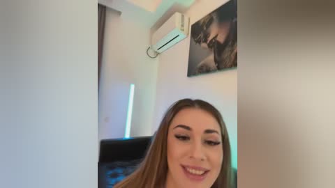 Media: A video of a smiling young woman with long, straight, light brown hair, wearing makeup and a black top, indoors. Background features a framed video, air conditioning unit, and a black sofa.