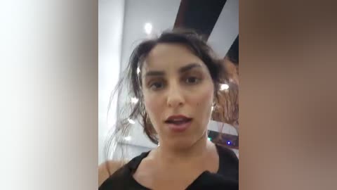 Video of a Latina woman with dark hair and olive skin, wearing a black top, speaking animatedly, indoors with modern decor.