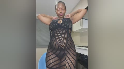 Video of a curvy, dark-skinned woman with short hair in a black, see-through mesh dress, showing off her ample cleavage and curves in a modern kitchen setting.