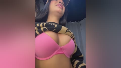 A video of a young woman with long black hair and medium skin tone, wearing a pink bra and a leopard print cardigan, standing in a dimly lit room.