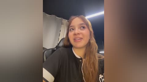Video of a young woman with long, light brown hair, wearing a black hoodie, smiling. She is seated in a dimly lit room with a dark ceiling and visible ceiling fan.
