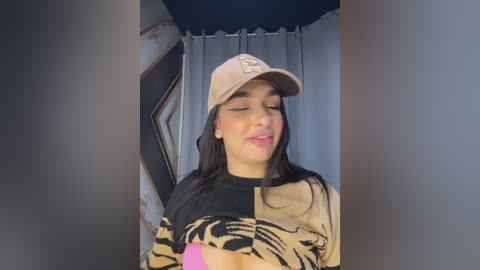 Video of a young woman with medium skin tone, long black hair, wearing a beige cap and a patterned sweater, smiling slightly, in a room with blue curtains and a wooden frame.