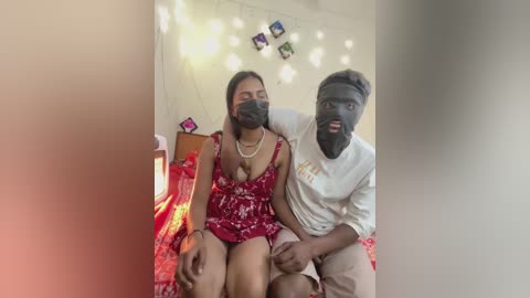 Video of a South Asian couple in a bedroom; the woman in a red dress, black face mask, and pearl necklace, sitting on the bed, the man in a white t-shirt, dark face paint, and shorts, with a surprised expression.