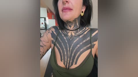 Video of a woman with medium skin tone, wearing a green tank top, showcasing intricate black tribal tattoos covering her chest, neck, and arms, with a nose ring, and short black hair.