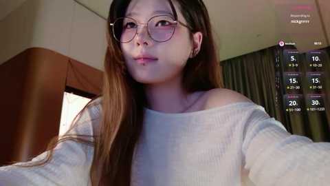Video of an East Asian woman with long brown hair, wearing round glasses, off-shoulder white sweater, and a neutral expression, indoors with dim lighting and a calendar overlay.