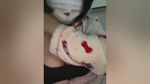 Video of a person wearing a white face mask, a red and black teddy bear onesie, and a black hoodie, lying on a bed with a pink blanket, blurred background.