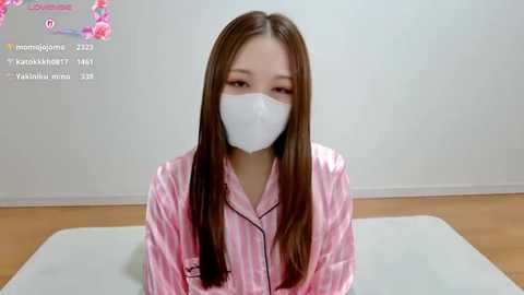 Media: Video of a young East Asian woman with long straight brown hair, wearing a pink and white striped pajama top and a white face mask, seated on a white table against a plain white wall.