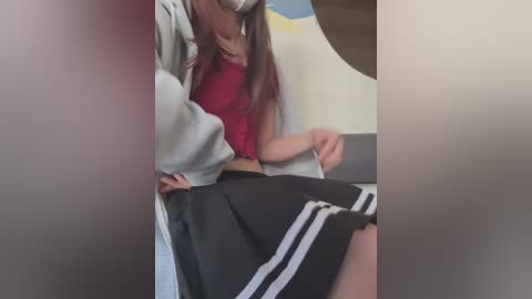 A video captures a young woman in a red top and black Adidas skirt, seated indoors, wearing a face mask.