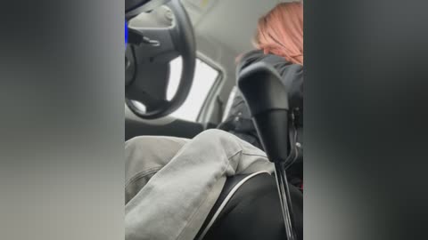 Video of a person wearing grey pants and a black jacket, seated in a car with a grey dashboard and steering wheel. The person's red hair is partially visible.