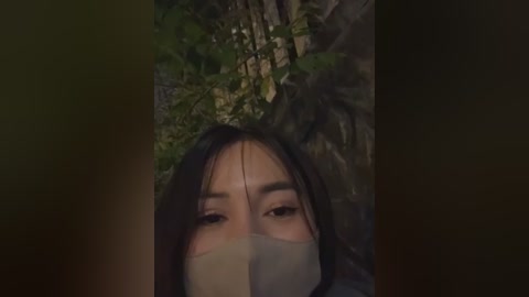 A video of a woman with long black hair, wearing a surgical mask, lying on a forest floor, surrounded by green foliage and dark shadows.