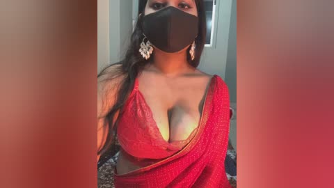 A video of a South Asian woman with medium skin tone, wearing a red saree, black face mask, and large silver earrings, with her breasts partially exposed due to the saree\u2019s cut.
