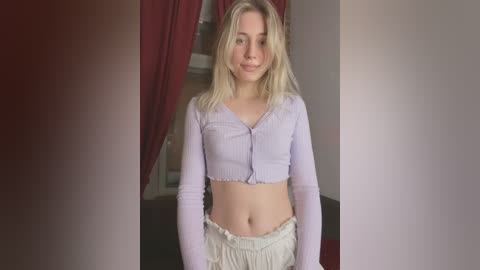 Video of a young blonde woman with fair skin, wearing a light purple ribbed crop top and white shorts, standing indoors with red curtains in the background.