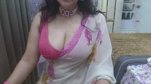 Media: Video of a woman with medium skin tone, dark hair, wearing a pink lace bra under a white sari with floral patterns, sitting indoors.