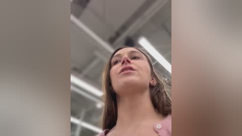 Media: A low-angle video of a young Caucasian woman with long brown hair, wearing a pink top, standing in a modern, well-lit room with visible ceiling beams and industrial-style lights.