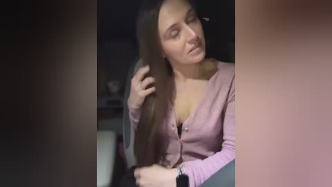 Media: A video of a young woman with long, straight brown hair, wearing a pink cardigan, seated indoors, looking relaxed.