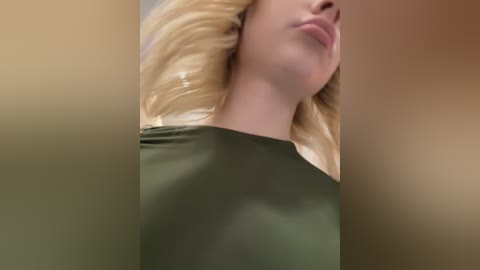 Video of a close-up of a blonde woman with light skin, wearing a green shirt, head tilted slightly, lips slightly parted. The background is a soft gradient of beige and brown.