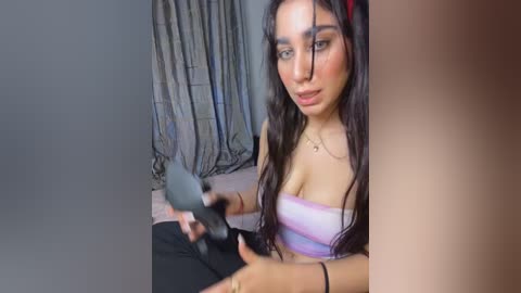 Video of a young woman with long dark hair, wearing a strapless purple top, sitting in a room with gray curtains, holding a makeup brush.